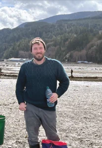 William Waller, owner of Living Stone Masonry, standing, smiling holding a water bottle in front of a mountain in the background with evergreen trees
