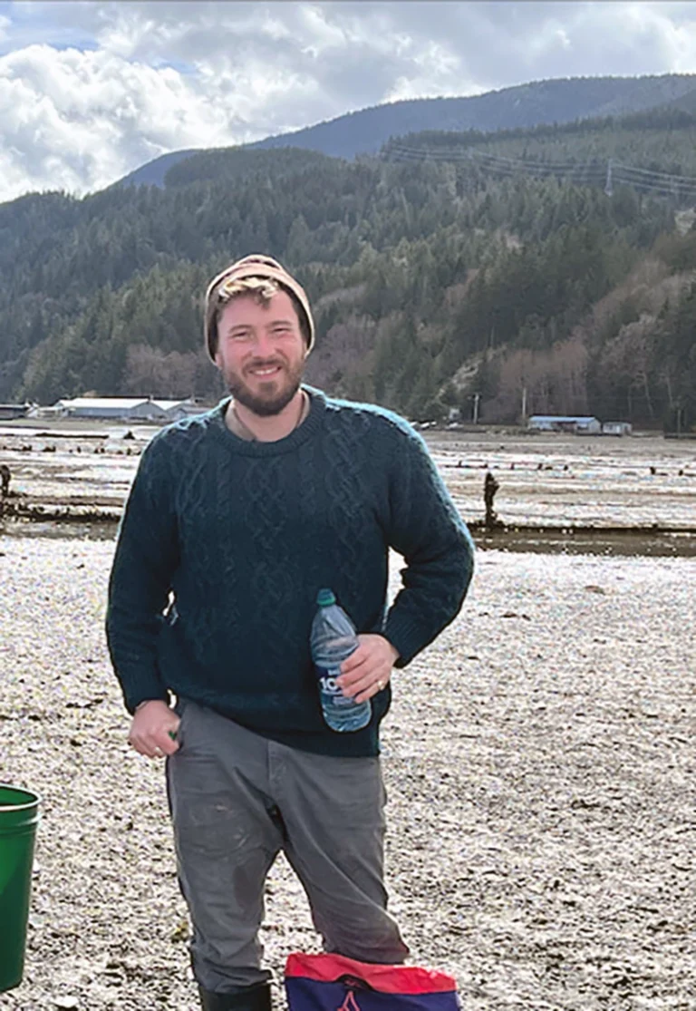 William Waller, owner of Living Stone Masonry, standing, smiling holding a water bottle in front of a mountain in the background with evergreen trees