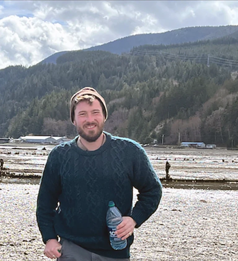 William Waller, owner of Living Stone Masonry, standing, smiling holding a water bottle in front of a mountain in the background with evergreen trees